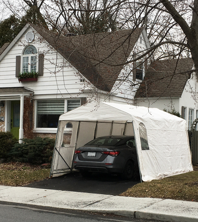 A grey car parked under a car shelter in the yard of a white house.