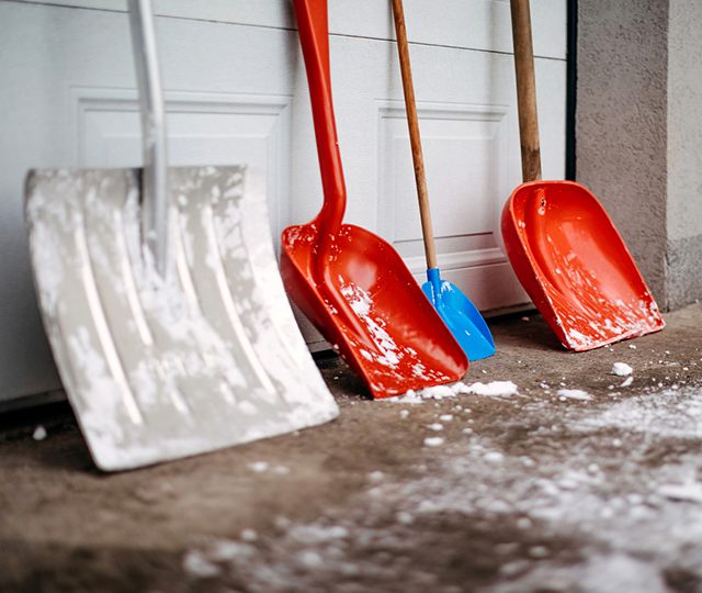 Plusieurs pelles à neige appuyées contre une porte de garage, dont des modèles rouges, bleus et une pelle métallique.