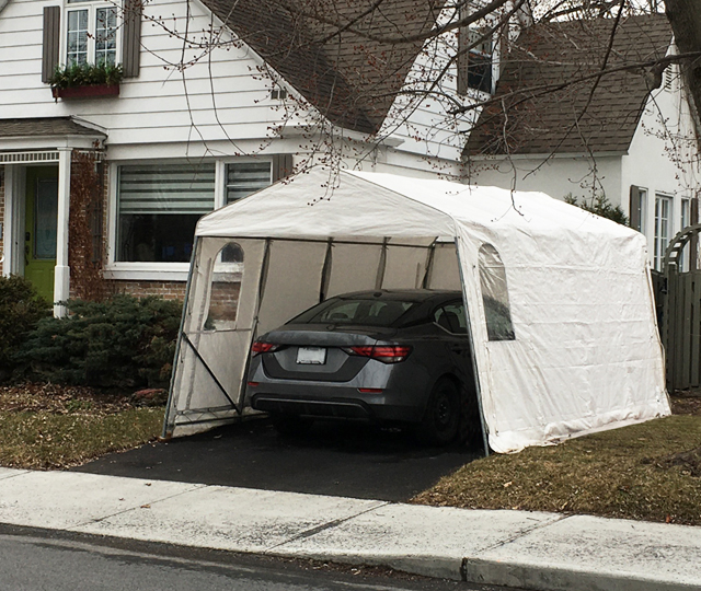A grey car parked under a car shelter in the yard of a white house.