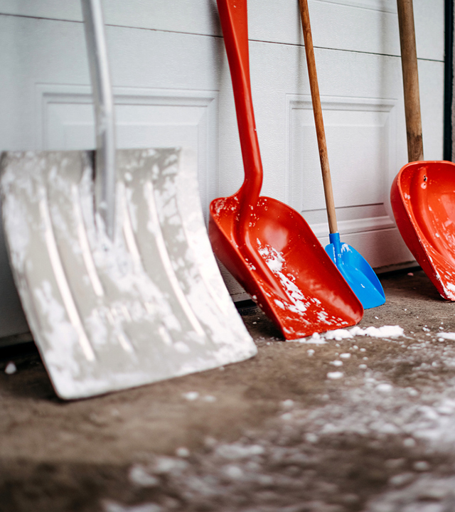 Snow shovels in various sizes leaning against a garage door, with snow residue on the ground.