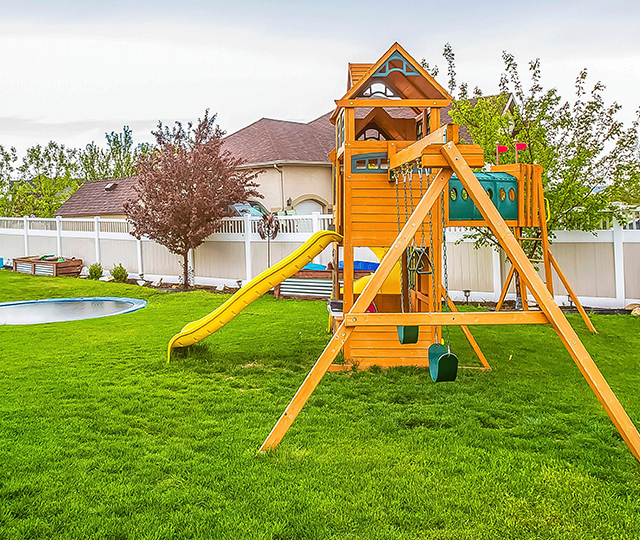 Cour arrière résidentielle avec une grande pelouse, un module de jeux en bois, un trampoline, des arbres et une clôture. Le visuel illustre les obstacles à repérer avant de tondre la pelouse pour travailler de façon sécuritaire.