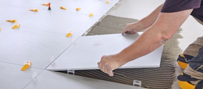 Gros plan sur un homme en train d'installer une tuile de céramique blanche sur un plancher dont quelques tuiles ont déjà été installées.