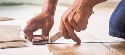 Très gros plan sur un homme en train de découper une planche de plancher SPC avec un exacto et une équerre.