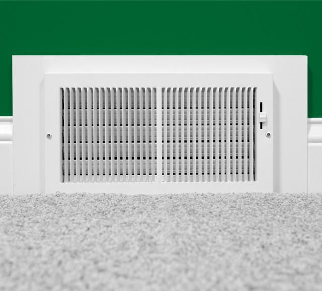 Grille de ventilation blanche d&rsquo;un syst&egrave;me de climatisation central install&eacute;e au bas d&rsquo;un mur vert, devant un tapis p&acirc;le.