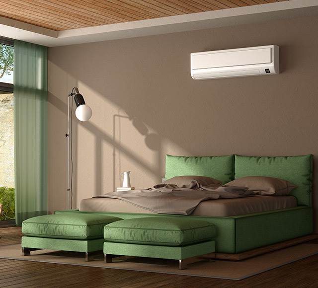 Climatiseur mural de type mini-split install&eacute; au-dessus d&rsquo;un lit dans une chambre moderne aux tons verts et bois.