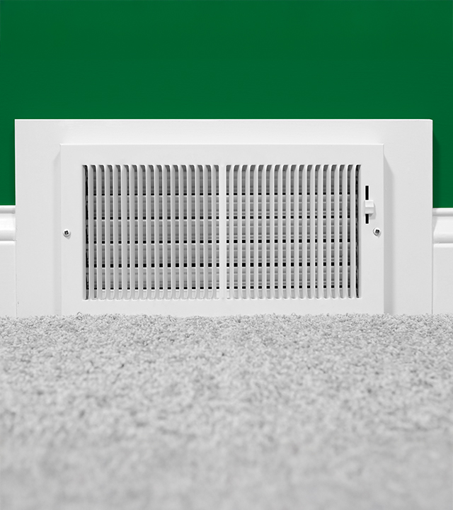 Grille de ventilation blanche d&rsquo;un syst&egrave;me de climatisation central install&eacute;e au bas d&rsquo;un mur vert, devant un tapis p&acirc;le.