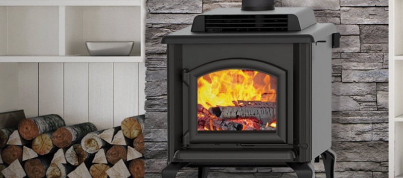 Black wood-burning stove with a lit fire, installed in front of a stone wall with firewood nearby.