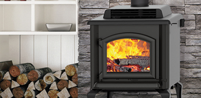Black wood-burning stove with a lit fire, installed in front of a stone wall with firewood nearby.
