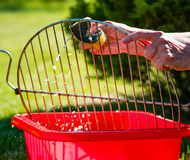 Nettoyage d&rsquo;une grille de barbecue avec une &eacute;ponge au-dessus d&rsquo;un bac d&rsquo;eau rouge &agrave; l&rsquo;ext&eacute;rieur.
