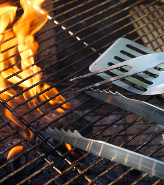 Grille de barbecue au charbon avec flammes visibles et ustensiles en acier pos&eacute;s dessus.