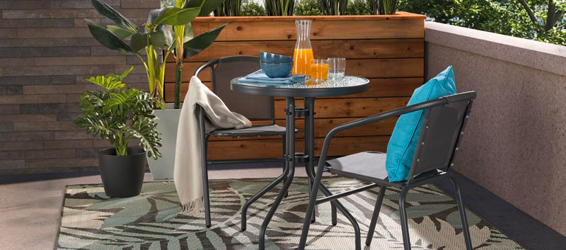 Small outdoor bistro set with round table and metal chairs on a balcony decorated with plants and an area rug.