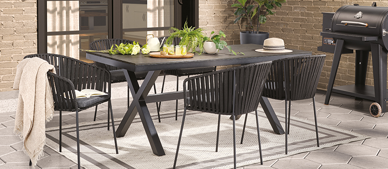 Outdoor dining set with a modern black table and chairs, set on an interlocking stone patio with a barbecue nearby.