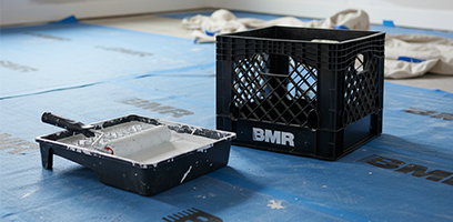 Bac à peinture avec rouleau et caisse noire BMR posés sur un plancher recouvert d’une membrane de protection bleue en préparation pour des travaux de peinture.