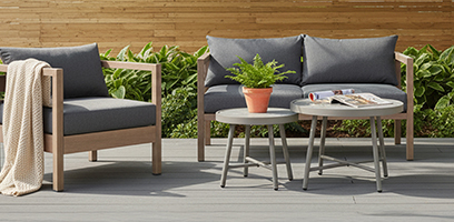 Ensemble de mobilier de patio moderne avec fauteuil et causeuse en bois clair et coussins gris, accompagn&eacute; de deux tables rondes et d&rsquo;une plante en pot, install&eacute; sur une terrasse ext&eacute;rieure.